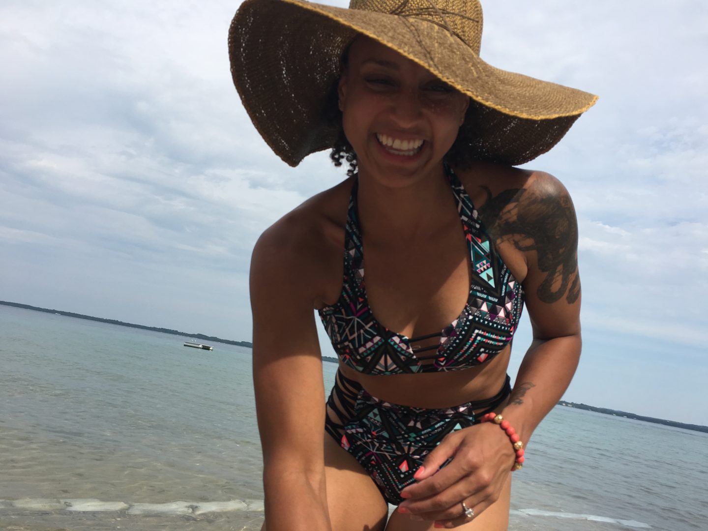 image of a woman at a beach, in a bathing suit and a big floppy hat, smiling at the camera