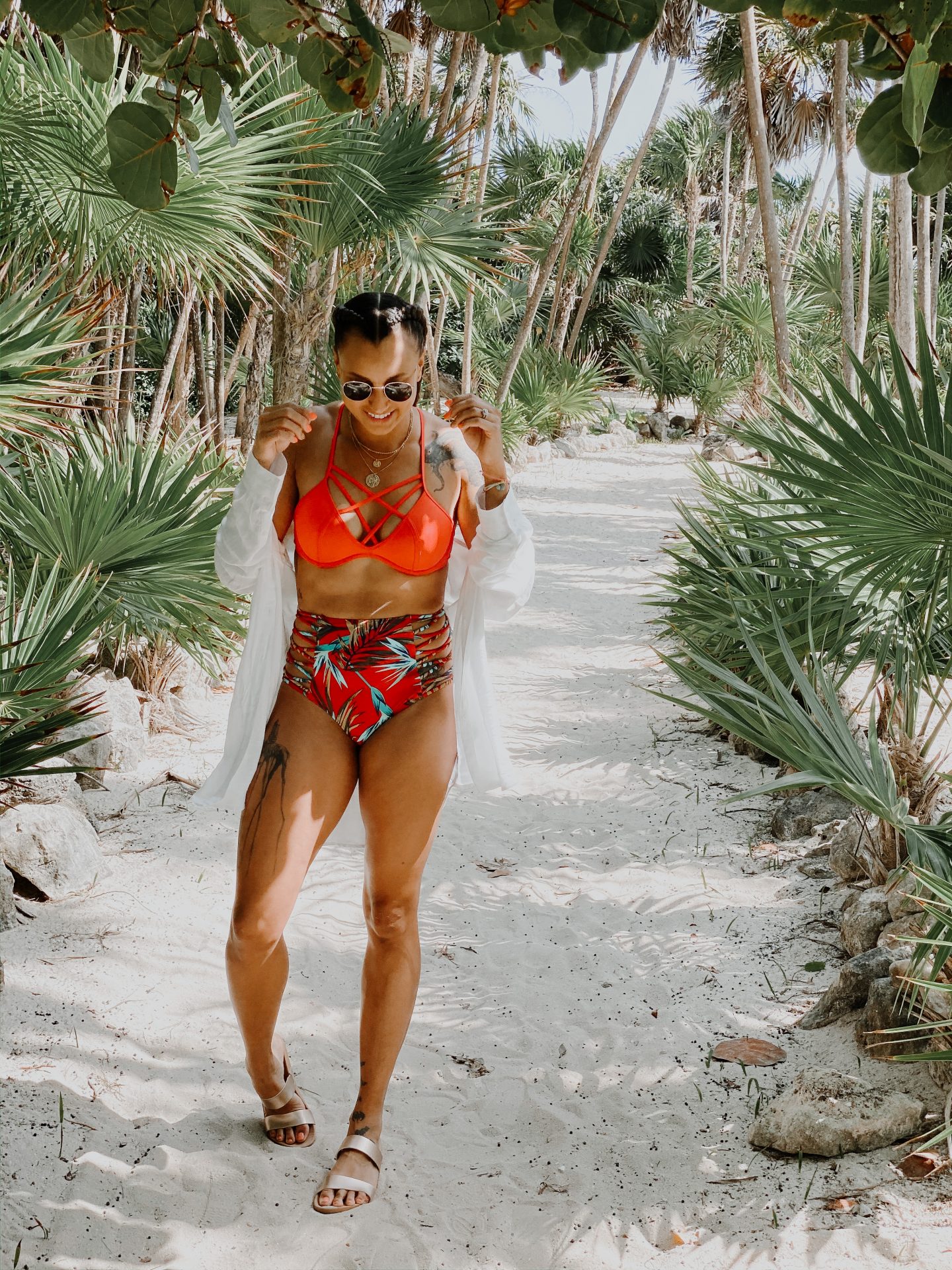 image of a mixed race woman, standing on a sandy path surrounded by palm trees and greenery. She is wearing a bright red, patterned two-piece swimsuit.