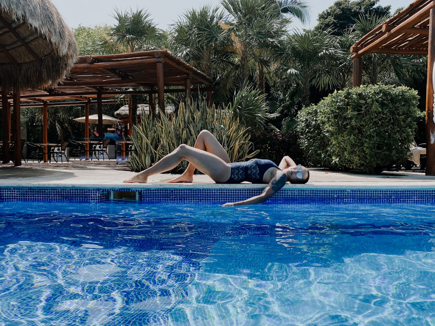 image of a mixed race female wearing a navy blue one-piece swimsuit, laying on the side of a very blue pool.