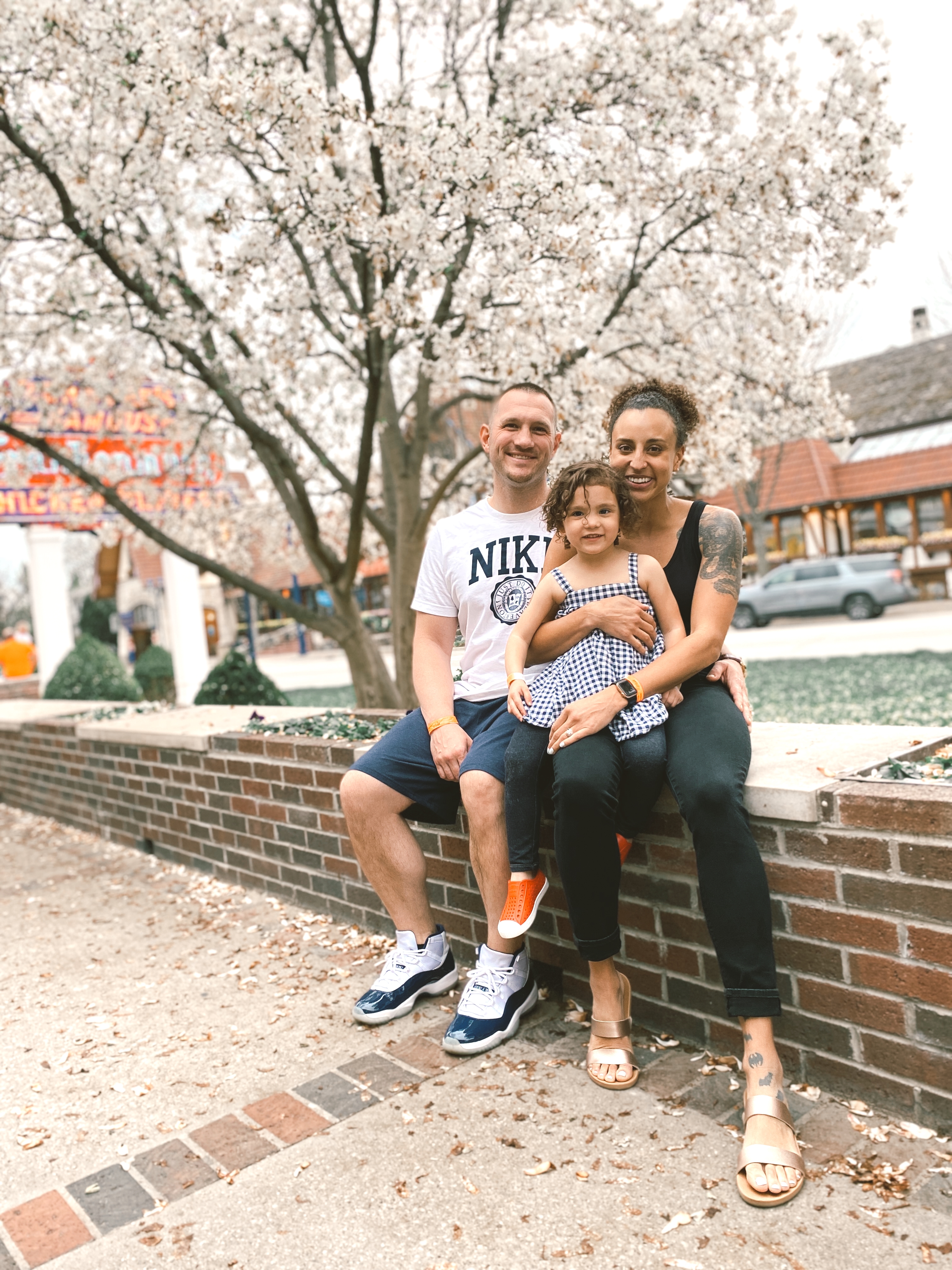 image of a family sitting on a brick ledge. There is a caucasian man, a mixed race woman, and a little girl. There is a big flowering tree in the background.